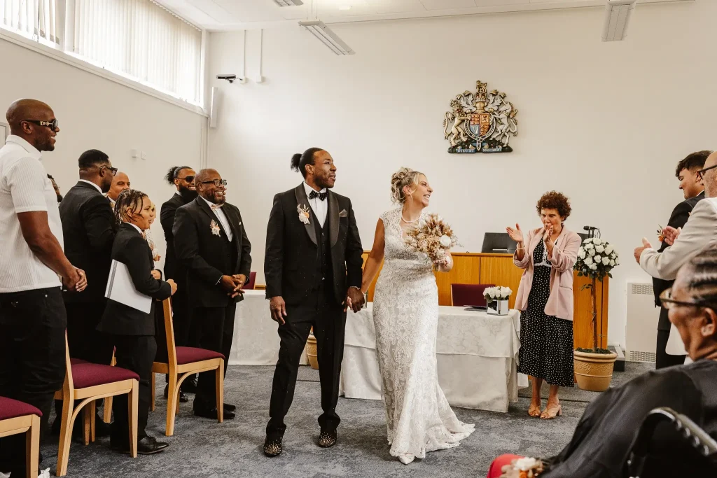 Newly married couple walking down the aisle at Beaconsfield Registry Office as family and friends stand and applaud following their wedding ceremony.