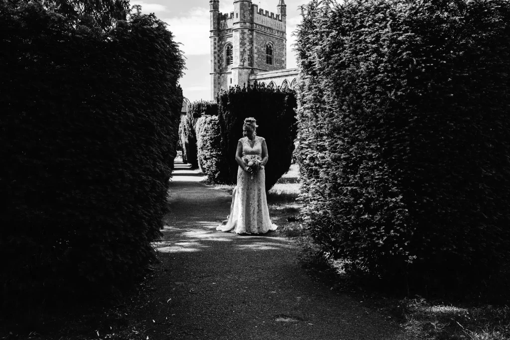 Bride standing in formal church gardens during a Buckinghamshire wedding, captured in a dramatic black and white documentary portrait.