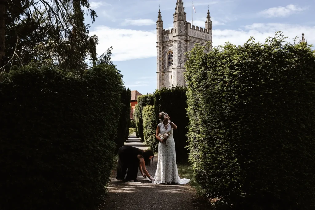 Bride standing in a formal garden pathway while a bridesmaid adjusts her wedding dress, with a historic church tower rising in the background.