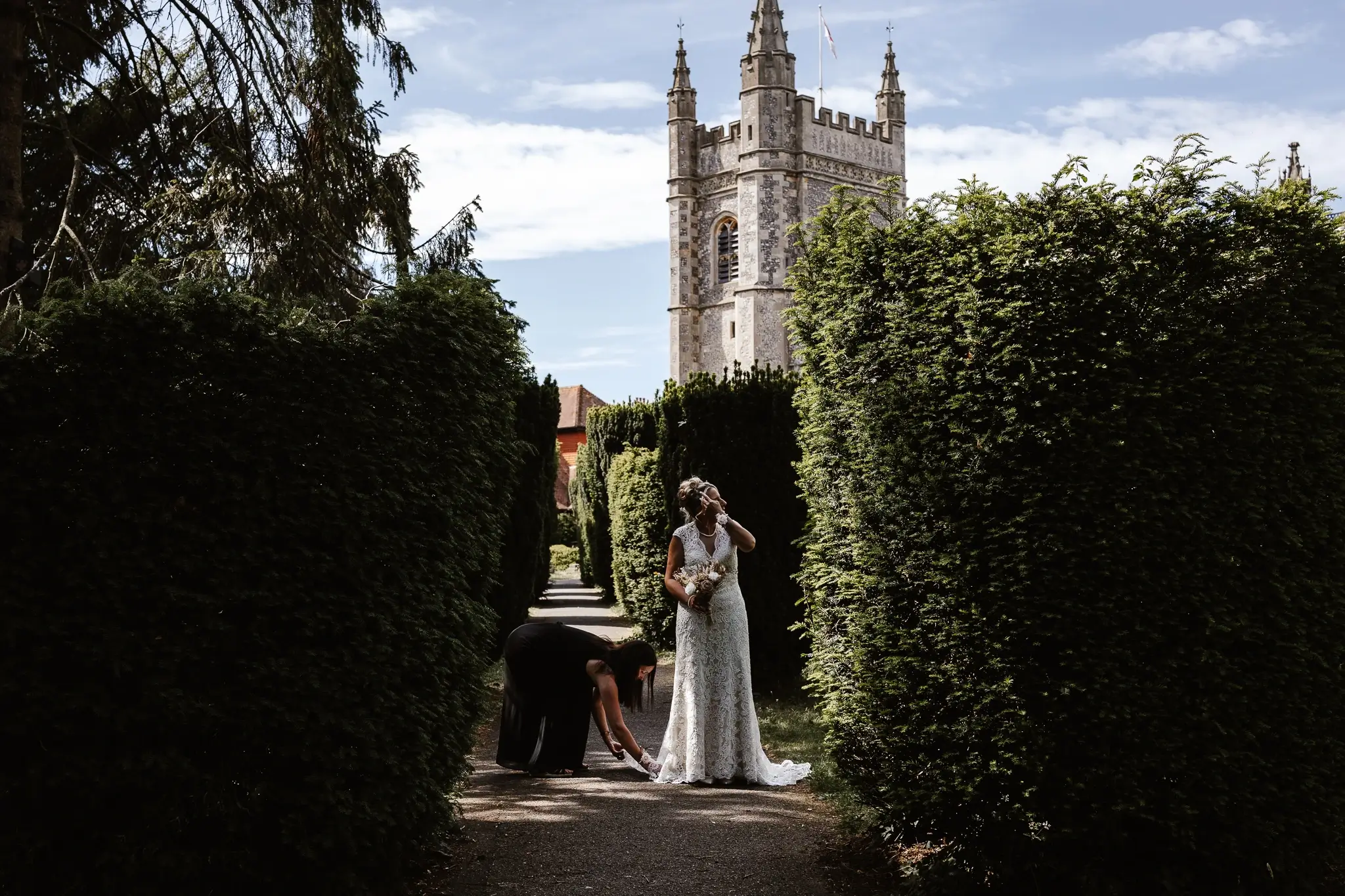 Bride standing in a formal garden pathway while a bridesmaid adjusts her wedding dress, with a historic church tower rising in the background.
